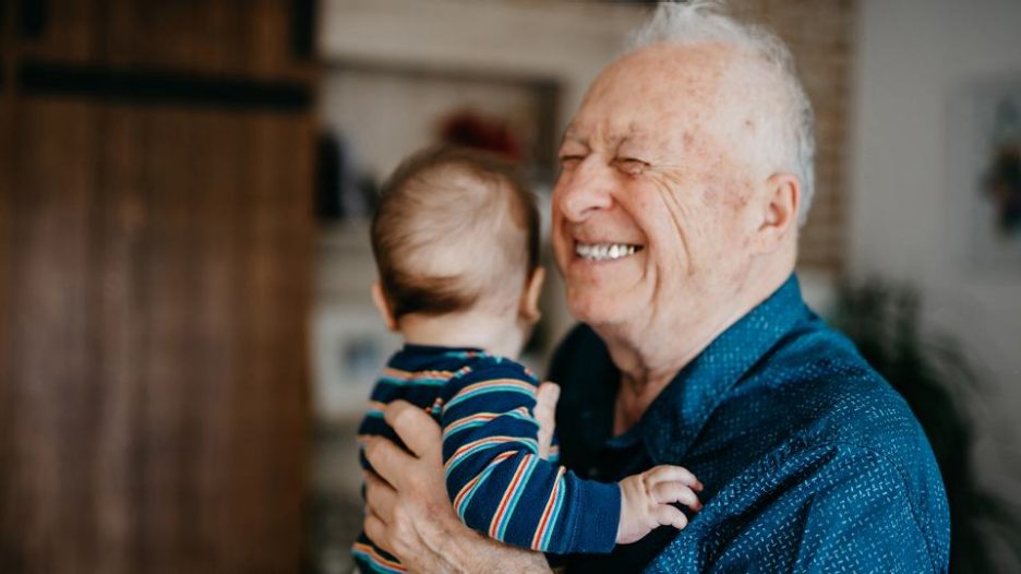 a smiling grandfather in a navy shirt is cuddling a baby inside